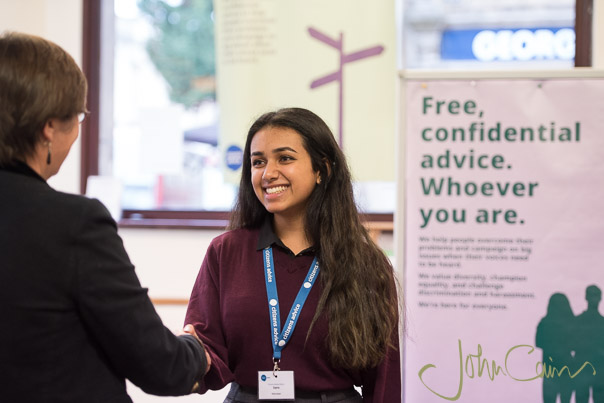An adviser welcomes a client, with a poster in the background saying "Free, confidential advice. Whoever you are."